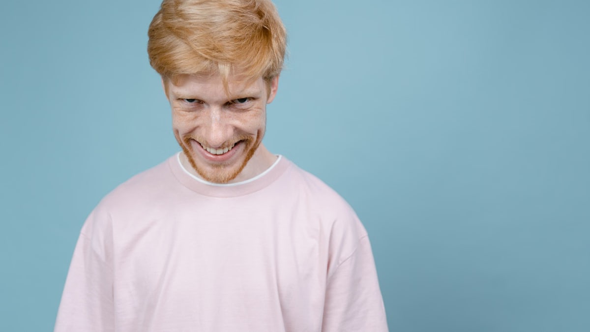 Young man in pink shirt smiles mischievously against blue background in studio setting.