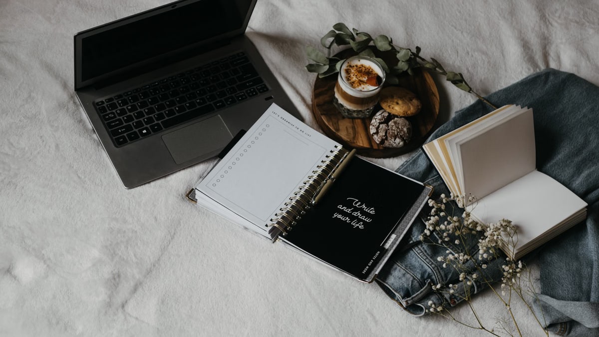 A cozy flat lay of a laptop, journals, and snacks on a bed for a relaxed work-from-home setup.